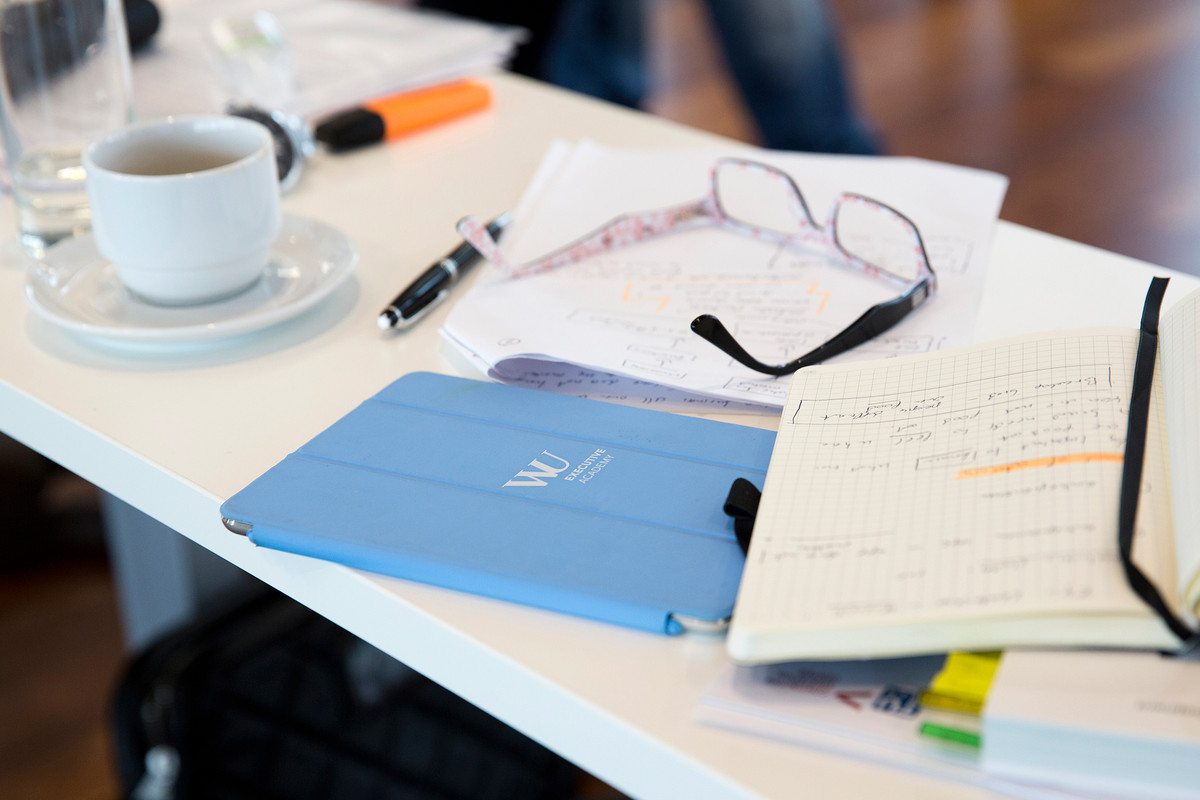 Arbeitsmaterialien, ein Tablet mit Logo der WU Executive Academy, eine Kaffeetasse, eine Brille, eine Füllfeder und ein Farbmarker auf einem Arbeitstisch