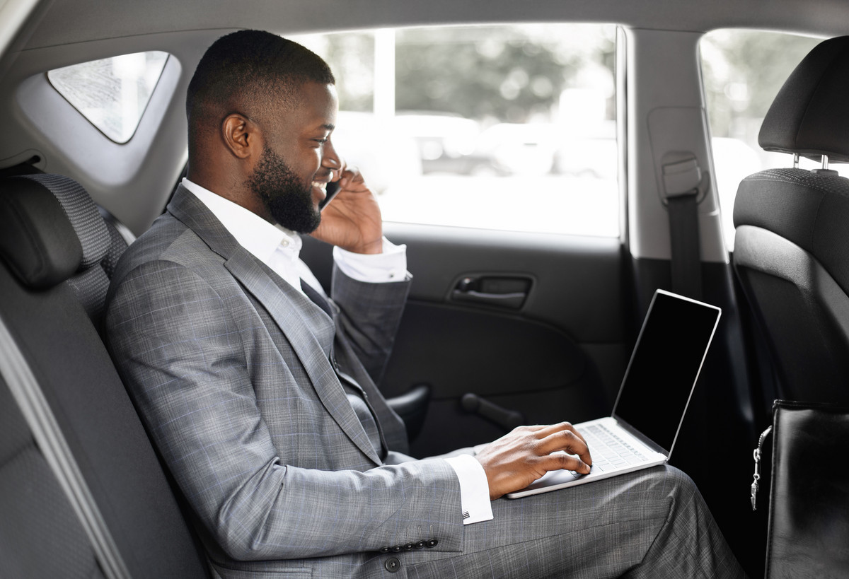 Ein Mann mit Mobiltelefon am Ohr und mit Laptop auf dem Schoß sitzt im Fond einer Limousine und telefoniert