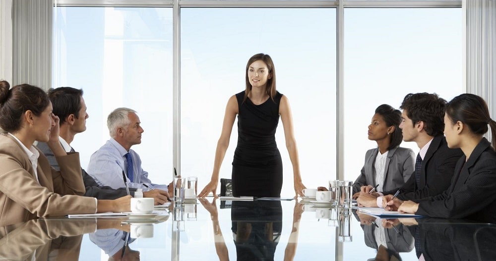 A woman stands at the head of a meeting table, many seated participants listen to her intently