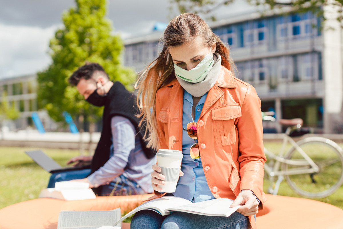 Eine junge Studentin mit Gesichtsmaske und Kaffeebecher in der Hand studiert ihr Lehrbuch, dahinter ein junger Mann, in seinen Laptop vertieft