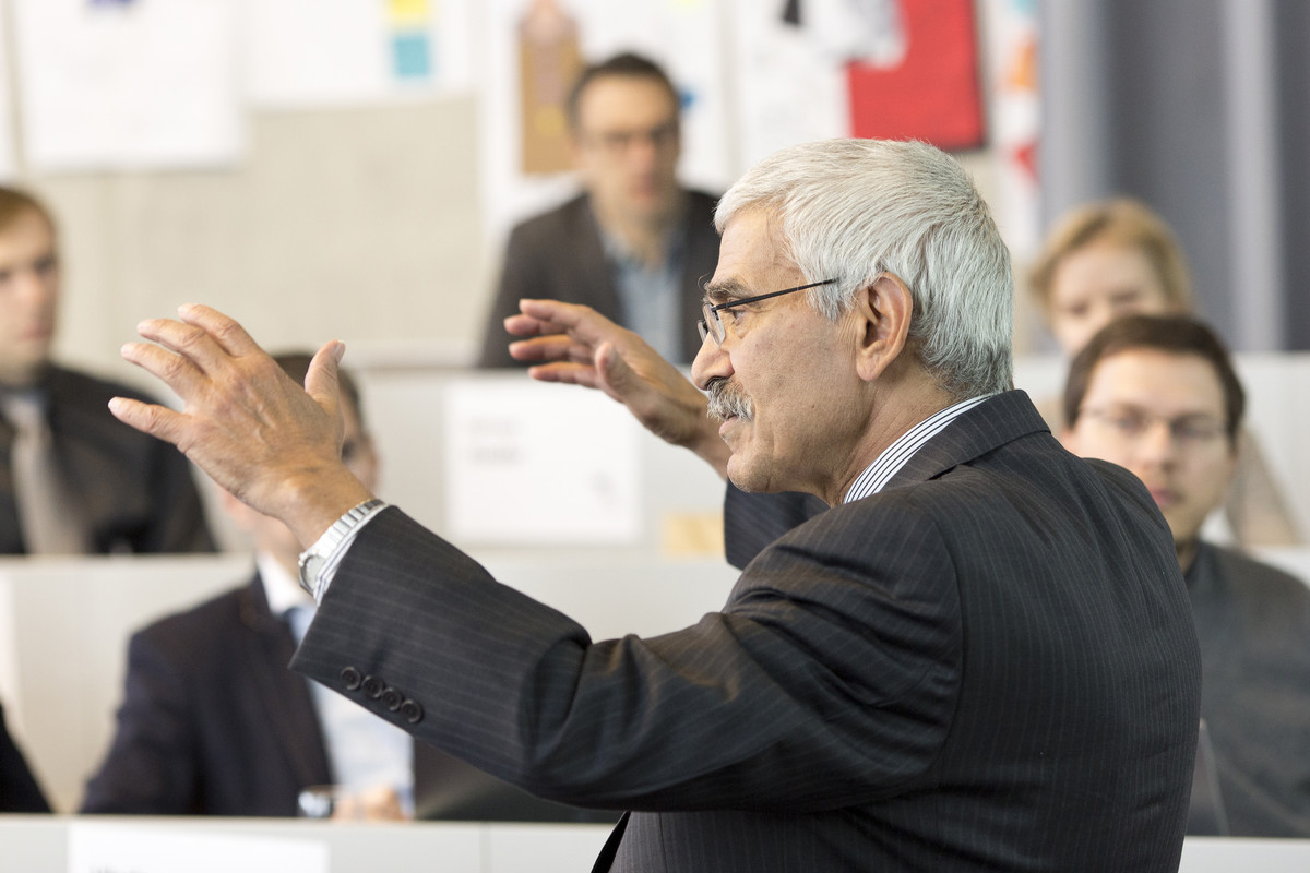 An elderly professor holding up both hands and giving a speech to an MBA class