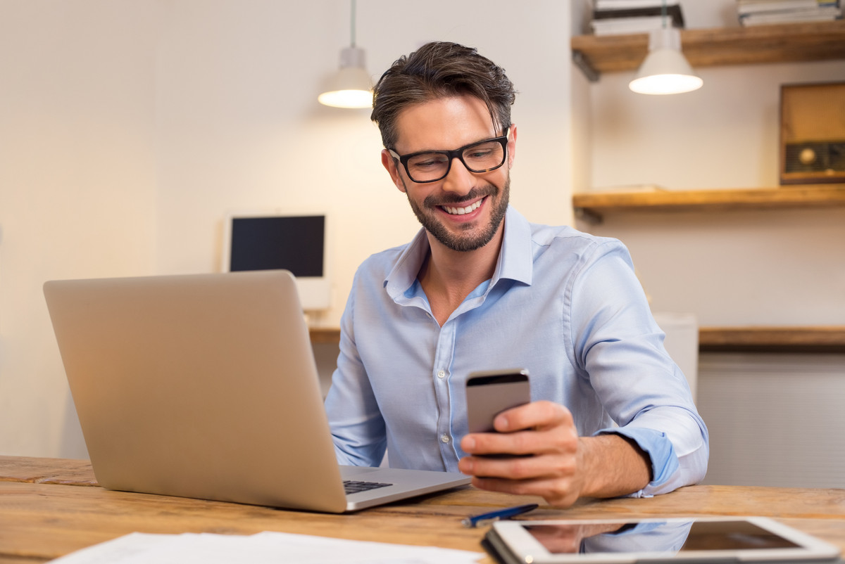 Young happy business man using smartphone in front of his laptop