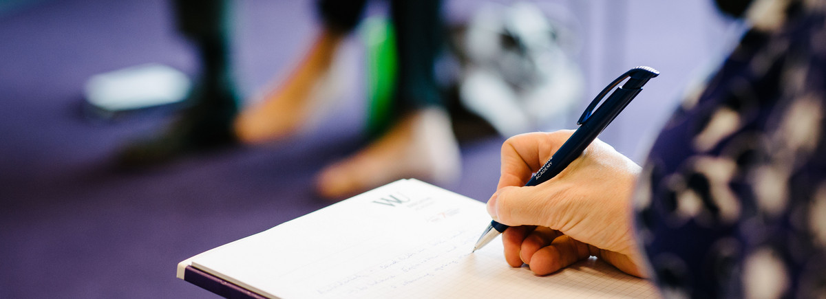 Eine weibliche Hand macht mit einem WU Executive Academy Kugelschreiber Notizen auf einem Block
