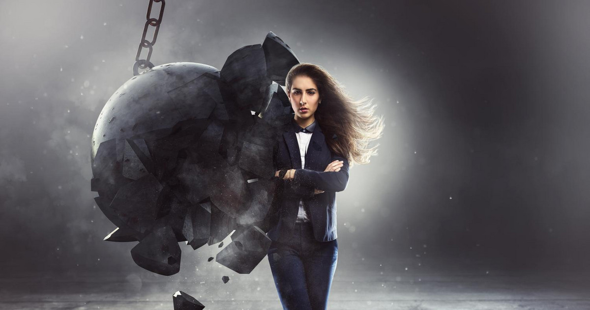 A woman stands confidently with her arms crossed, a wrecking ball shatters against her coming from the left side