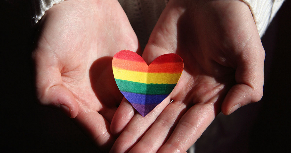 Two hands hold a heart made of paper in rainbow colors