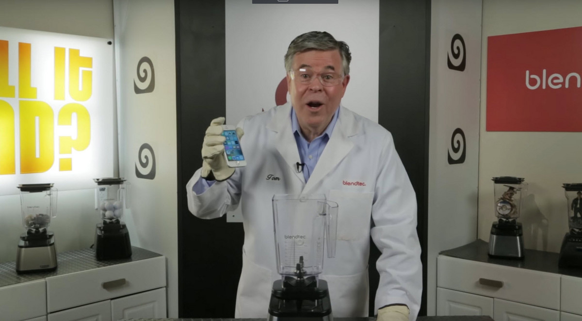 A man in a white science suit holding an iPhone above a blender