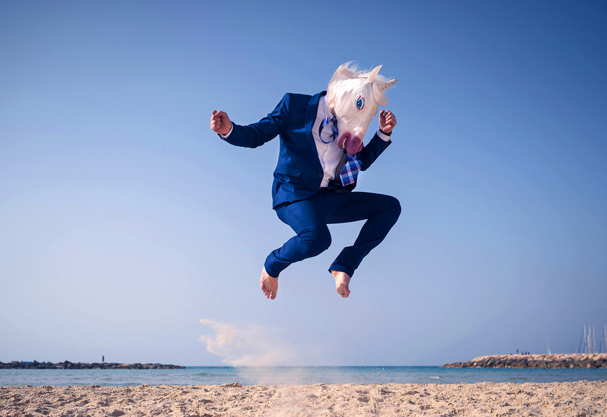 Ein Mann im Anzug und mit einer Einhörnmaske spirngt am Strand in die Luft