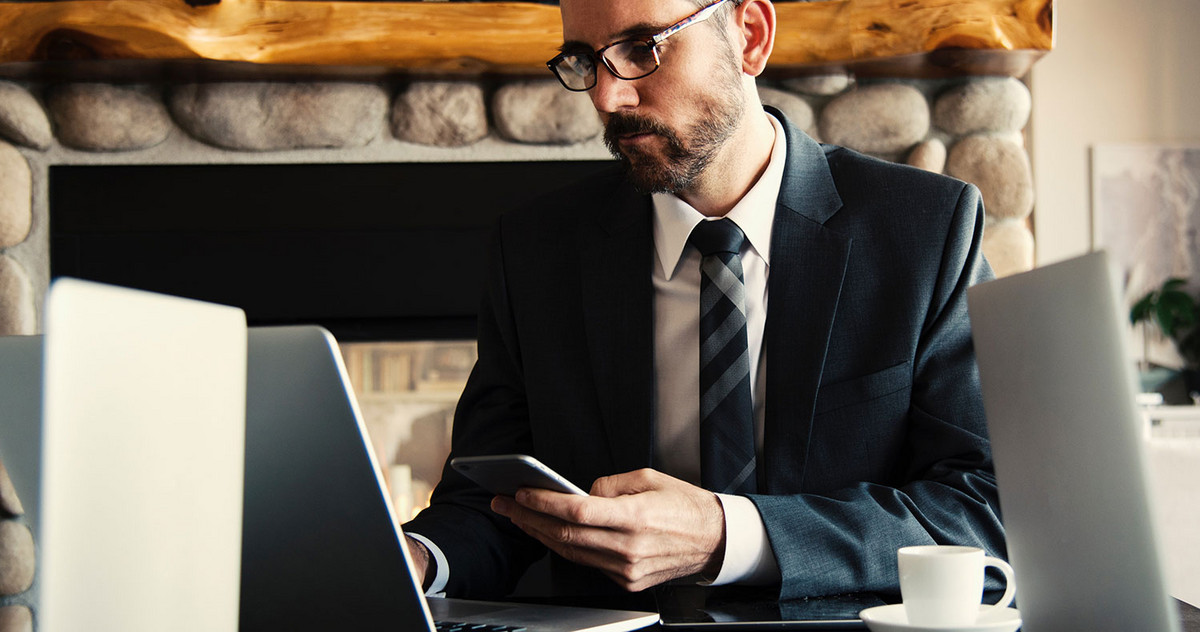 Ein Mann im Anzug mit Smartphone in der linken Hand arbeitet auf seinem Laptop