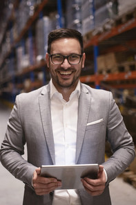 Ein Mann im Anzug, mit einem Tablet in der Hand in einer Lagerhalle vor vollen Regalen