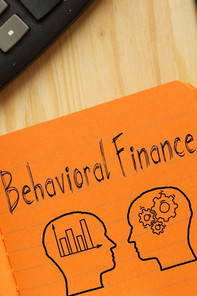 A calculator and a notepad with "Behavioral Finance" written on it, with a drawing of two heads below it