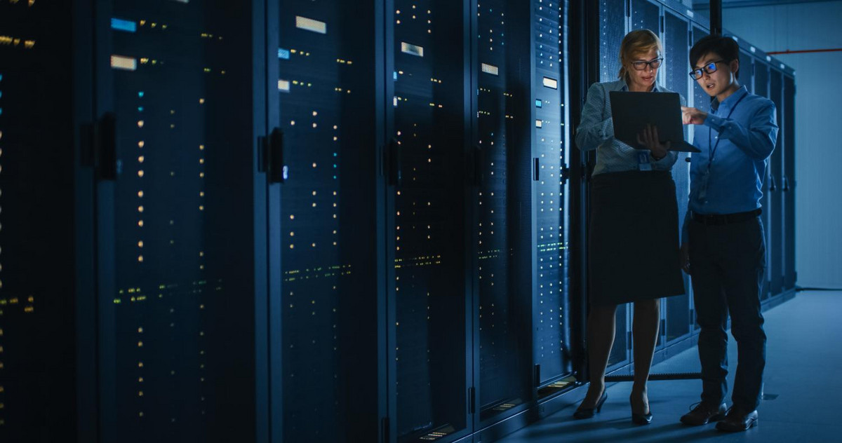 A woman holding a laptop and a man pointing at the laptop are standing in a server room