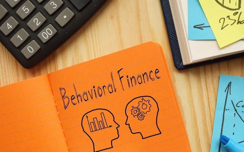 A calculator and a notepad with "Behavioral Finance" written on it, with a drawing of two heads below it